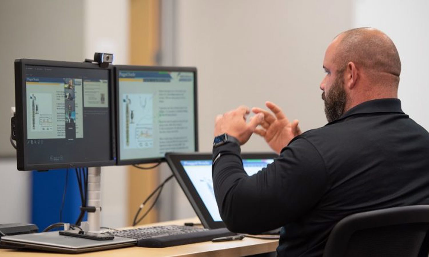 A person sitting at a desk with dual monitors and a tablet, gesturing with their hands, possibly during an online meeting or presentation.