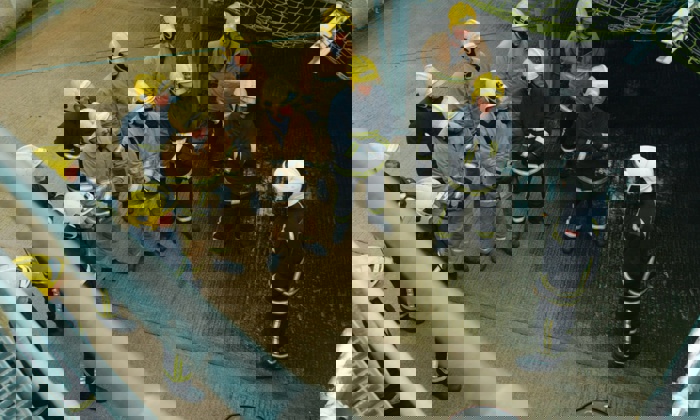 A group of firefighters dressed in yellow helmets and firefighting gear stands in formation, listening to another firefighter in white helmet giving instructions.