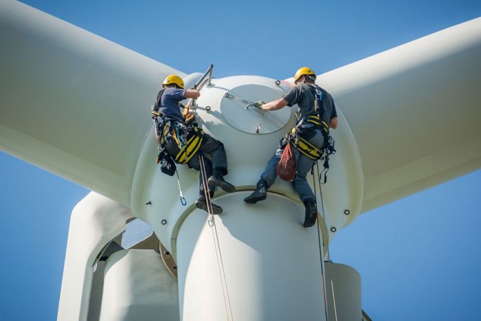 Two workers in safety harnesses and helmets are performing maintenance at the top of a wind turbine against a clear blue sky.
