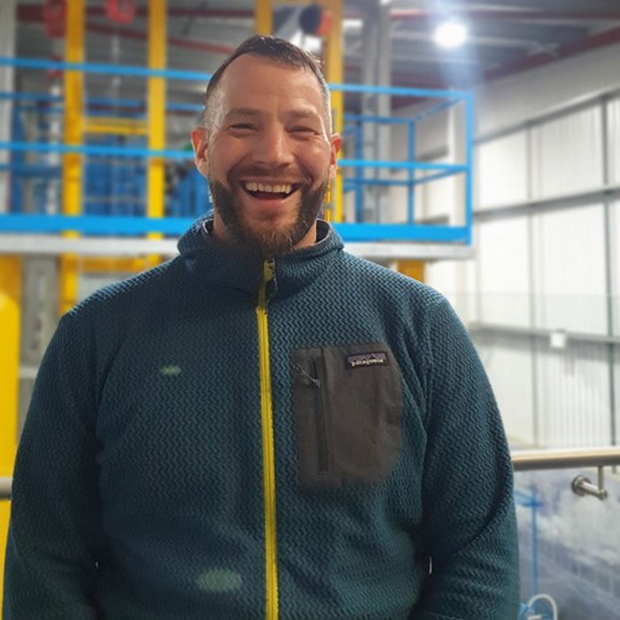 A man in a blue fleece jacket stands indoors in front of industrial equipment, smiling broadly at the camera.