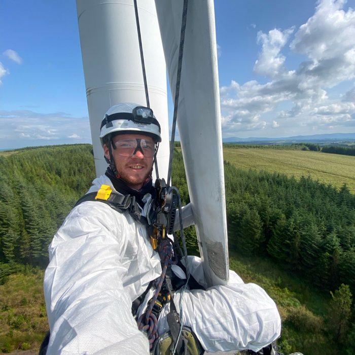 A person in protective gear and helmet is suspended by ropes on a wind turbine, with a forested landscape and clear sky in the background.