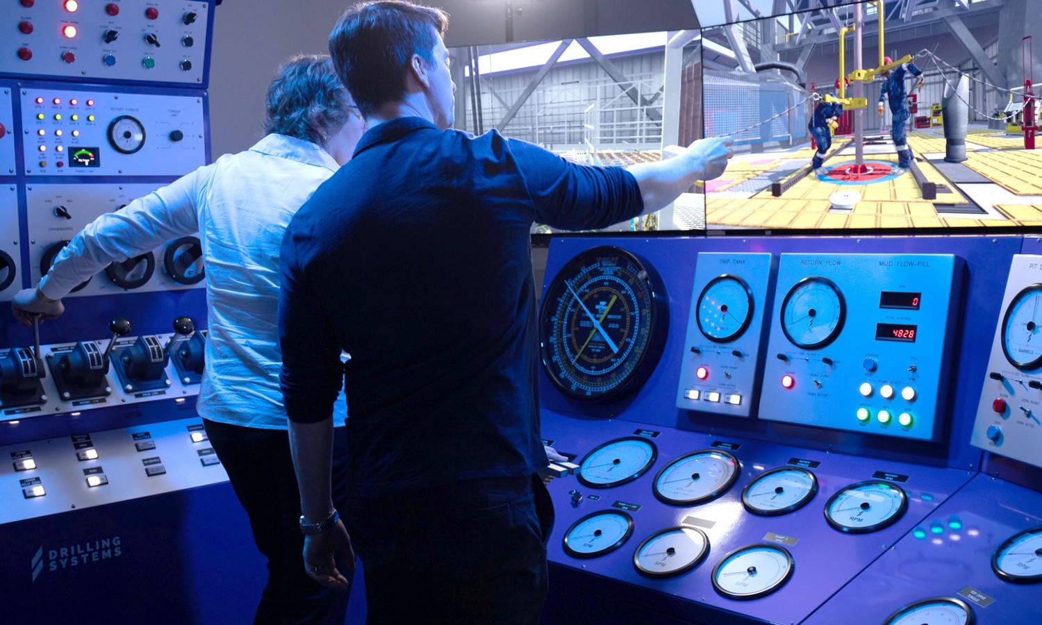 Two individuals operate a control panel in a drilling simulation room, with gauges and screens displaying real-time drilling activities.