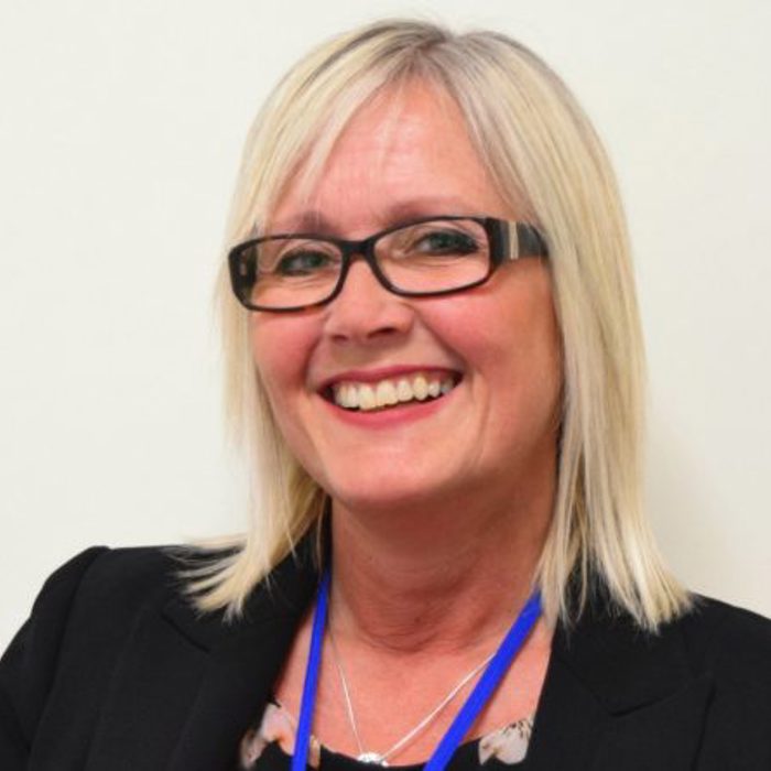A smiling woman with blonde hair and glasses, wearing a black blazer with a blue lanyard, stands against a plain background.