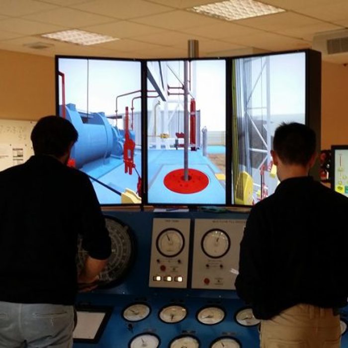 Two people operating a simulator with multiple screens and control panels in a control room setting.