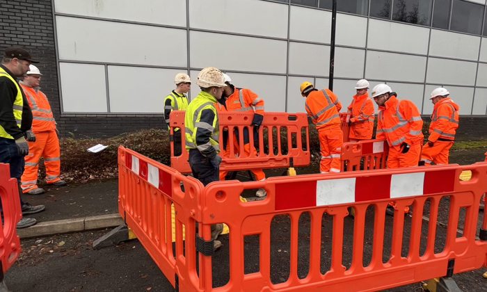 Construction workers in high-visibility clothing and hard hats gather around orange safety barriers on a street next to a building, demonstrating their expertise from comprehensive utilities and construction training.