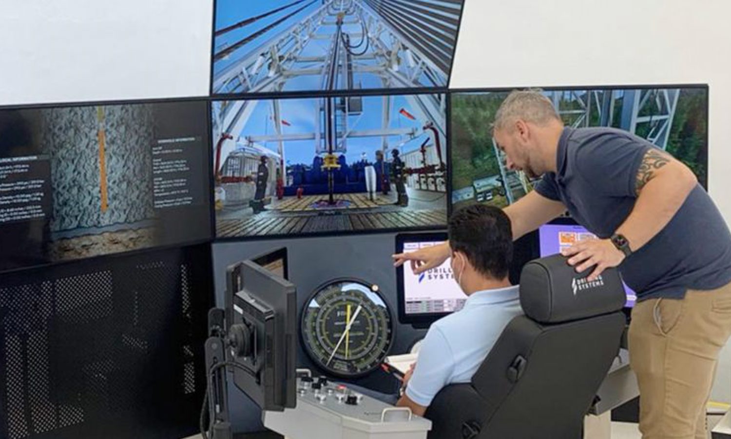 Two men in a control room monitor multiple screens displaying drilling operations. One seated man operates controls while another man stands beside him, pointing to a screen.