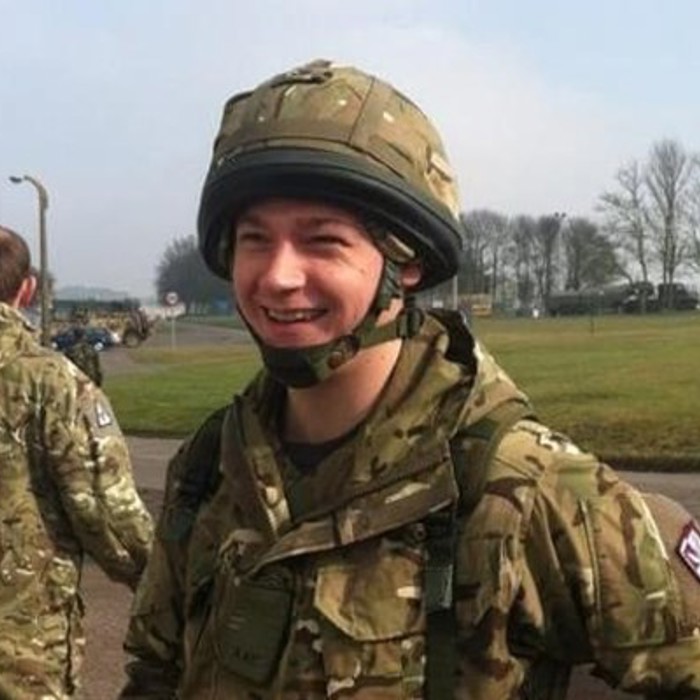 A person in military uniform smiles outdoors near a building and a parked motorcycle, with other uniformed individuals and trees in the background.