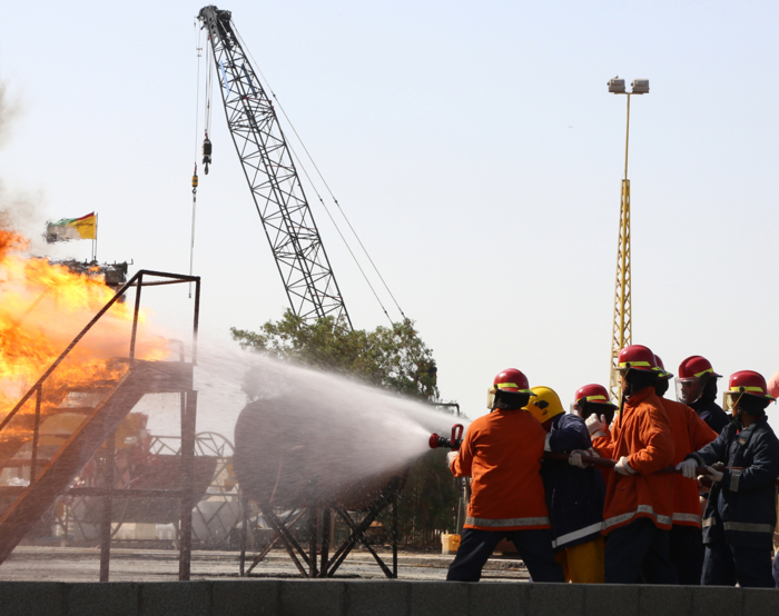 Firefighters in orange uniforms and red helmets use a hose to spray water on a large industrial fire with thick smoke. A crane and flag are visible in the background.