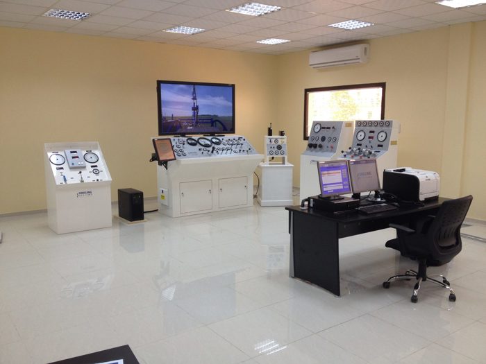 A control room with multiple control panels, a desk with computer monitors, various gauges, and a large screen on the wall displaying an image of an industrial setup.