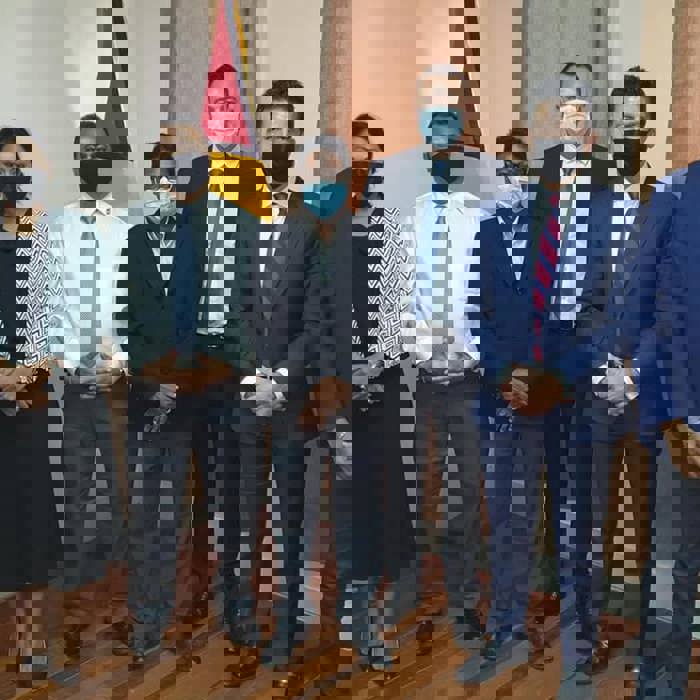 A group of seven people, wearing formal attire and face masks, stand in a room with curtains and a flag in the background.