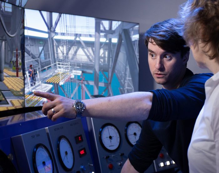 A person points at a screen displaying an industrial setting while explaining something to another person. Dials and gauges are visible on a control panel in the foreground.