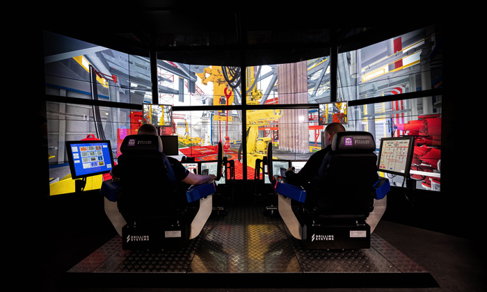 Two individuals are seated at a simulator displaying a virtual construction site with cranes and equipment on multiple screens. In front of them are control panels and a small touchscreen.