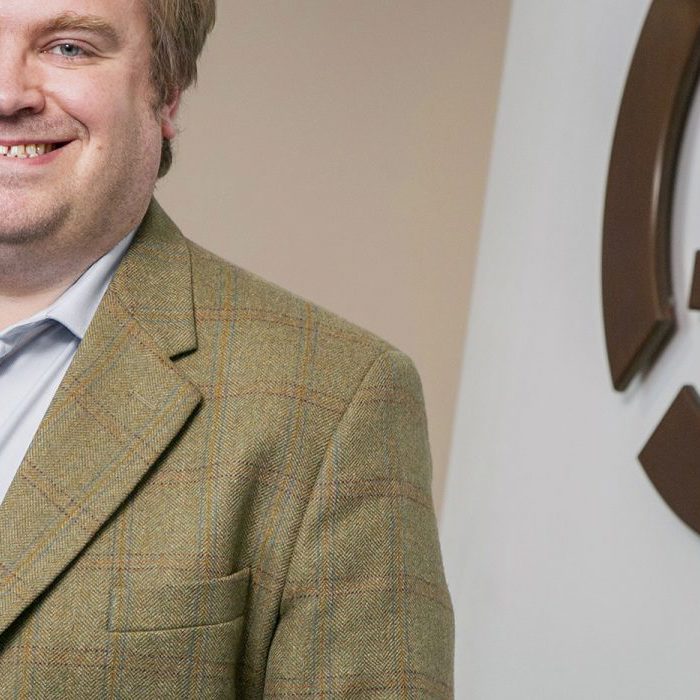 A man in a plaid blazer and light blue shirt stands smiling next to a wall with a partial logo featuring a stylized 'S'.